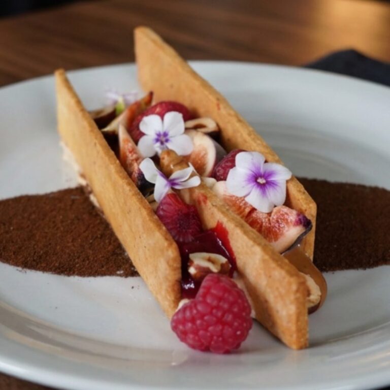 Dessert à l'assiette : biscuit garni de figues, framboises et fleurs comestibles, sur un trait de poudre.
